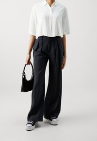 Chemise blanche raccourcie à boutons avec manches courtes ; pantalons amples bleu marine à plis ; sac à main noir ; baskets noires et blanches.