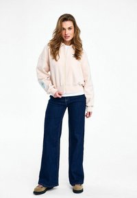 Žena s dlhými vlasmi stojaca, oblečená v bledo ružovej bunde na zips, tmavých širokých džínsach a hnedých šnurovacích topánkach na bielom pozadí.