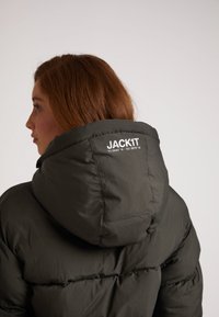 Giacca imbottita verde scuro con un grande cappuccio. Presenta un logo sul retro e una trama trapuntata e gonfi. Materiale liscio e resistente all'acqua.