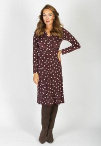Kobieta z długimi falującymi włosami, ubrana w bordową sukienkę w kropki i brązowe kozaki do kolan, stoi z jedną ręką na biodrze.