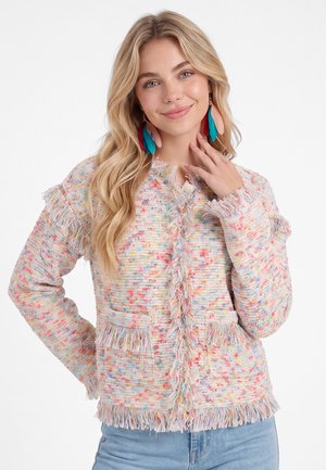 Junge Frau mit langen blonden Haaren, die eine bunte Fransenjacke und große rote, blaue und rosa Federohrringe trägt, lächelt mit der Hand nahe am Gesicht.