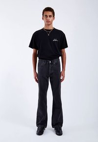 Black cotton t-shirt with front logo and black denim jeans with a flared design, worn with black shoes. Plain background, standing pose.