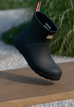 Black matte Hunter rain boot with red-white-black pull tab, suspended above wooden surface, green foliage blurred in background.