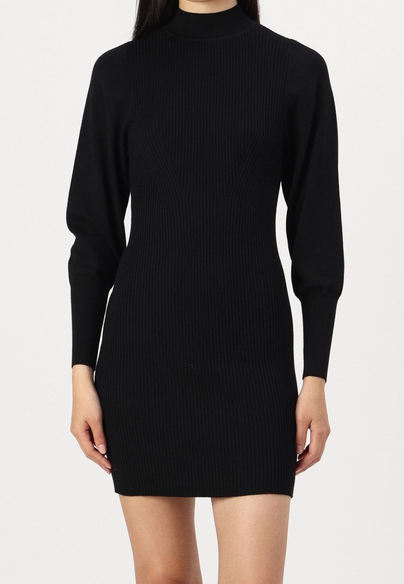 Femme portant une robe noire ajustée en maille côtelée avec manches longues et col montant, se tenant devant un fond clair uni.