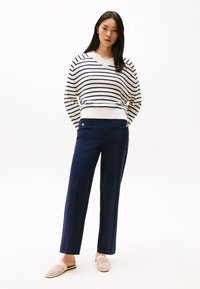 Pull rayé crème et marine, avec un col en V ; pantalon marine avec poches avant et boutons dorés ; chaussures à enfiler tissées de couleur claire.