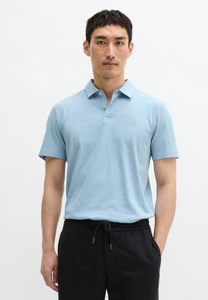 Mężczyzna w jasnoniebieskiej koszulce polo z krótkim rękawem włożonej do czarnych spodni ze sznurkiem, stojący na jednolitym białym tle.