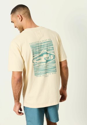 Mann trägt ein beigefarbenes T-Shirt mit blauem Fischgrafik und Text "Freestyle Surf Co. From Reef to Ride", kombiniert mit blauen Shorts, stehend vor einem einfarbigen Hintergrund.