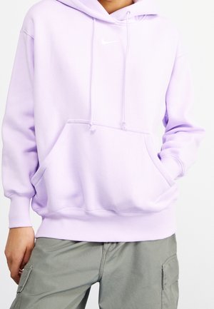 Persoon in een lichtpaarse hoodie met een voorzak en een wit Nike-logo, gecombineerd met grijze cargobroeken, met de hand naast zich rustend.