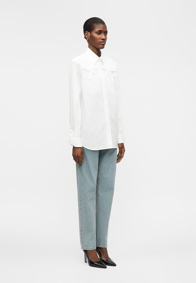 Femme debout portant une chemise blanche ample à manches longues, un jean bleu clair droit et des chaussures noires à talons hauts pointus.