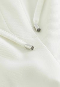 Witte, gestructureerde stof van een hoodie met twee zilveren metalen aglets aan de trekkoorden, die een schone en minimalistische uitstraling laat zien.