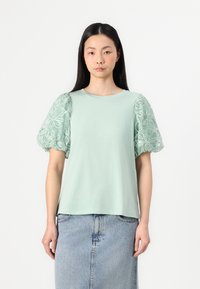 Gaiši zaļš T-kreklis ar puffatām mežģīņu piedurknēm, kas rotātas ar izšūtiem ziedu rakstiem. T-krekla apkakle ir apaļa, un tas ir brīva piegriezuma.