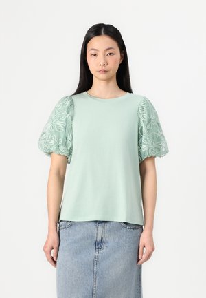 T-shirt verde claro com mangas de renda bufantes, apresentando padrões florais bordados. A camisa tem um decote redondo e um ajuste descontraído.