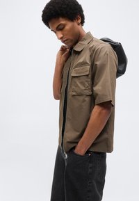 Νέος άνδρας με μαύρο αφροκρά hairstyle φοράει ελιά κοντό μανίκι πουκάμισο με φερμουάρ, μαύρα τζιν, κρατώντας μαύρο σακίδιο στην πλάτη ενάντια σε λευκό φόντο.