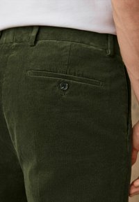 Ciemnozielone spodnie kordrowe z pionowym żebrowaniem, tylną kieszenią z zapięciem na guzik oraz delikatnie teksturowaną powierzchnią.