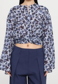 Blouse boutonnée en tissu léger, présentant un motif floral bleu et foncé, design court, et manches longues légèrement évasées.