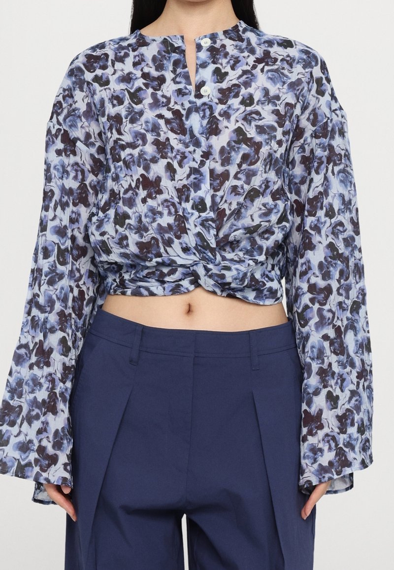 Blouse boutonnée en tissu léger, présentant un motif floral bleu et foncé, design court, et manches longues légèrement évasées.