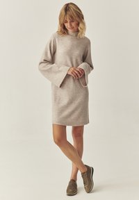 Fonalból készült, világos bézs színű ruha, magas nyakkal és bővülő ujjakkal. Barna sportcipőkkel párosítva, hogy laza megjelenést kölcsönözzön.