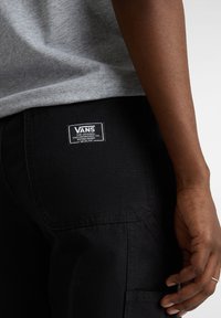 Zwarte broek van een gestructureerde stof, met een achterzak en een witte Vans-logo label. De achtergrond is neutraal en onopdringerig.