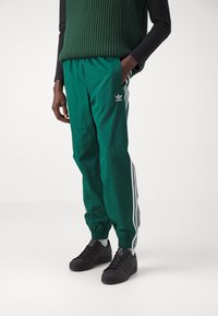 Zelené tepláky s bílými bočními pruhy, pružný pas a logo Adidas na levé stehenní části; v kombinaci s černými teniskami.