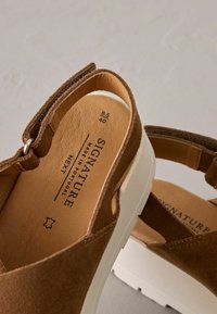 Sandali in suede marrone con punta chiusa e retro aperto, dotati di una suola bianca, logo impresso e un design di cinturini semplice.