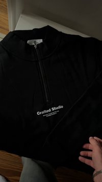 Sweatshirt preto com fecho de correr até à altura do peito, com o texto "Crafted Studio" no peito, colocado de forma plana sobre uma superfície; mão com um bracelete a tocar na mangueira do lado direito.