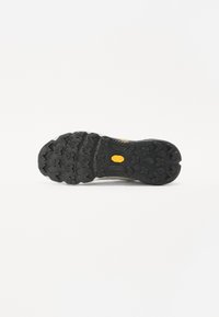 Semelle de chaussure noire en caoutchouc avec des motifs de traction profonds et un logo jaune visible. Surface texturée avec des formes variées pour une meilleure adhérence.