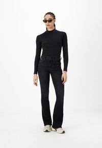 Black long-sleeve fitted top paired with dark denim flared jeans. Model wears sunglasses and light-colored sneakers. Plain white background.