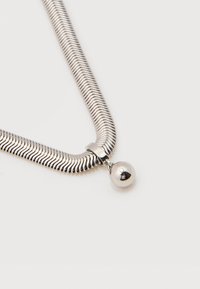 Collier en chaîne serpent argentée avec un pendentif rond et lisse en argent suspendu au centre sur un fond blanc uni.