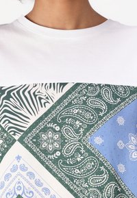 Chemise blanche à manches courtes avec un design en patchwork comprenant des imprimés animaliers et des motifs cachemire en vert, bleu et blanc. Matériau en coton.