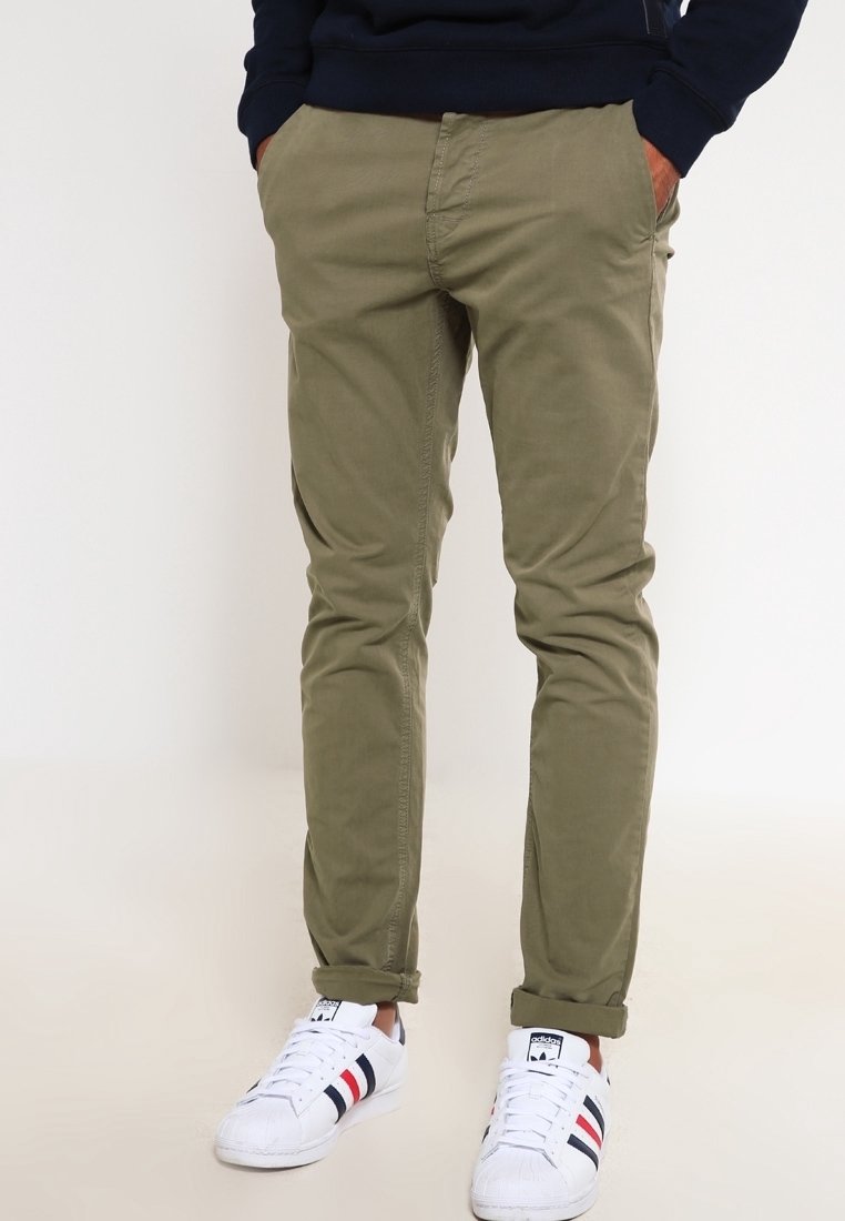 Homme portant un pantalon slim vert olive aux ourlets retroussés et des baskets blanches Adidas avec des rayures noires et rouges, les mains dans les poches.