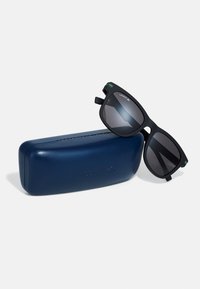 Ochelari de soare negri cu lentile întunecate, cu un finisaj mat și dungi verzi de accent pe ramă, așezați pe o carcasă rigidă albastră.