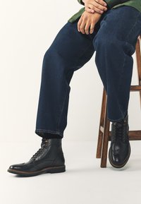 Schwarze Lederstiefel mit Brogue-Details, rundem Zehenbereich und Schnürverschluss. Braune Außensohle mit Ziernähten. Kombiniert mit dunkelblauen Jeans.