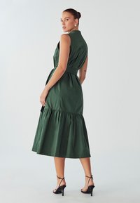 Roheline, varrukateta kleit, millel on vöödiga talje ja kihiline äär. Valmistatud siledast kangast, kõrge krae ja mustade plätudega.