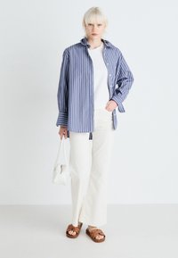 Košile s dlouhým rukávem modro-bílé pruhované, přes bílé tričko, v kombinaci s krémovými širokými kalhotami a hnědými pantoflemi, držící bílou tašku.