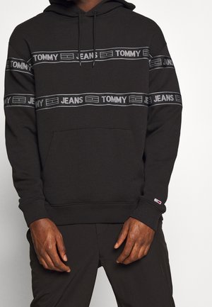 Musta huppari, jossa on "TOMMY JEANS"-teksti ja logoviirut rinnassa ja hihoissa, henkilön yllä, jonka kädet näkyvät vyötäröllä.