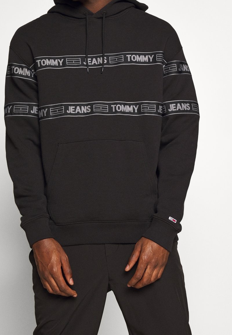 Sweat à capuche noir avec le texte « TOMMY JEANS » et des bandes logo sur la poitrine et les manches, porté par une personne avec les mains visibles à la taille.