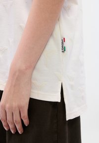 Λευκό βαμβακερό t-shirt με υφασμένο αφηρημένο μοτίβο, πλευρικές σχισμές στο τελείωμα και μικρή πολύχρωμη ετικέτα στην αριστερή πλευρά.