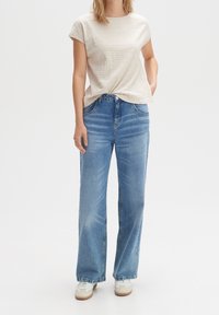 Nainen, joka on pukeutunut vaaleanbeigeen lyhythihaiseen neulepaitaan, korkeavyötäröisiin vaaleansinisiin suoriin farkkuihin ja valkoisiin lenkkareihin valkoista taustaa vasten.