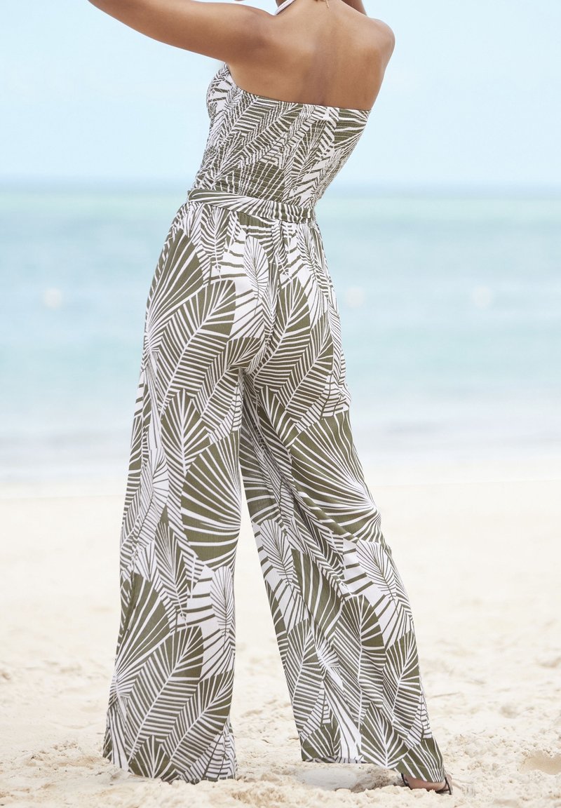 Femme portant une combinaison bustier avec un motif de feuilles vertes et blanches, debout sur une plage de sable près d'une mer calme.