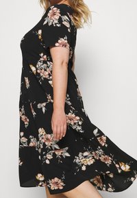 Musta kukallinen mekko, jossa on lyhyet hihat, pyöreä kaula-aukko ja väljää helmaa, jossa on vaaleanpunaisia ja kultaisia kukkakuoseja. Kevyt kangas.