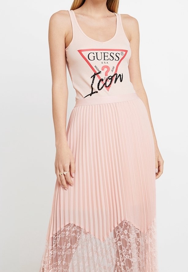 Femme portant un haut sans manches Guess rose pâle avec logo et une jupe plissée assortie avec ourlet en dentelle.