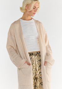 Blondynka nosząca beżowy sweter z dzianiny na białej koszuli i spodniami w wężowy wzór z wysokim stanem, z rękami w kieszeniach swetra.