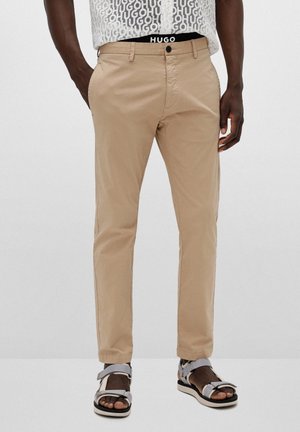Homme portant un pantalon chino beige, des sandales blanches et une chemise blanche avec un motif répété du logo "HUGO".