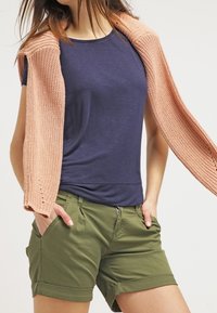 Navyblauw shirt met korte mouwen gecombineerd met een licht perzikkleurige gebreide cardigan en groene shorts met omgeslagen mouwen en zijzakken.