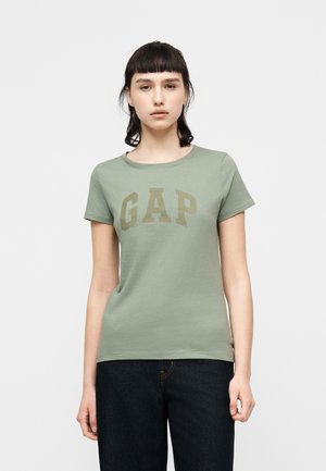 Junge Frau mit kurzen dunklen Haaren, die ein hellgrünes GAP-T-Shirt und dunkelblaue Jeans trägt, vor einem schlichten hellen Hintergrund stehend.