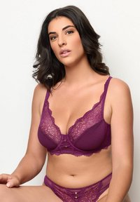 Žena s tmavými vlasy, oblečená v fialové krajkové podprsence a odpovídajících krajkových kalhotkách, sedí na jednotném bílém pozadí.