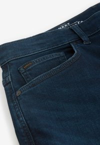 Blugi din denim de culoare albastru închis cu o buzunar frontal care prezintă un buzunar utilitar cusut. Cusăturile distincte și un mic eticheta cu logo sunt vizibile.