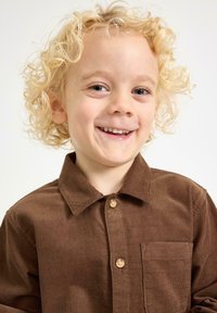 Brązowa koszula z sztruksu z kołnierzykiem, z kieszonką i drewnianymi guzikami, noszona przez dziecko z kręconymi blond włosami na neutralnym tle.
