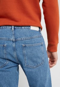 Jasnoniebieskie dżinsy z denimu o klasycznym kroju, z dwoma tylnymi kieszeniami i białą metką na pasku, noszone z pomarańczowym swetrem.