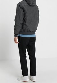 Giacca bomber con cappuccio grigio scuro sopra una camicia azzurra chiara, abbinata a pantaloni neri affusolati e sneakers bianche, presentando un outfit casual.